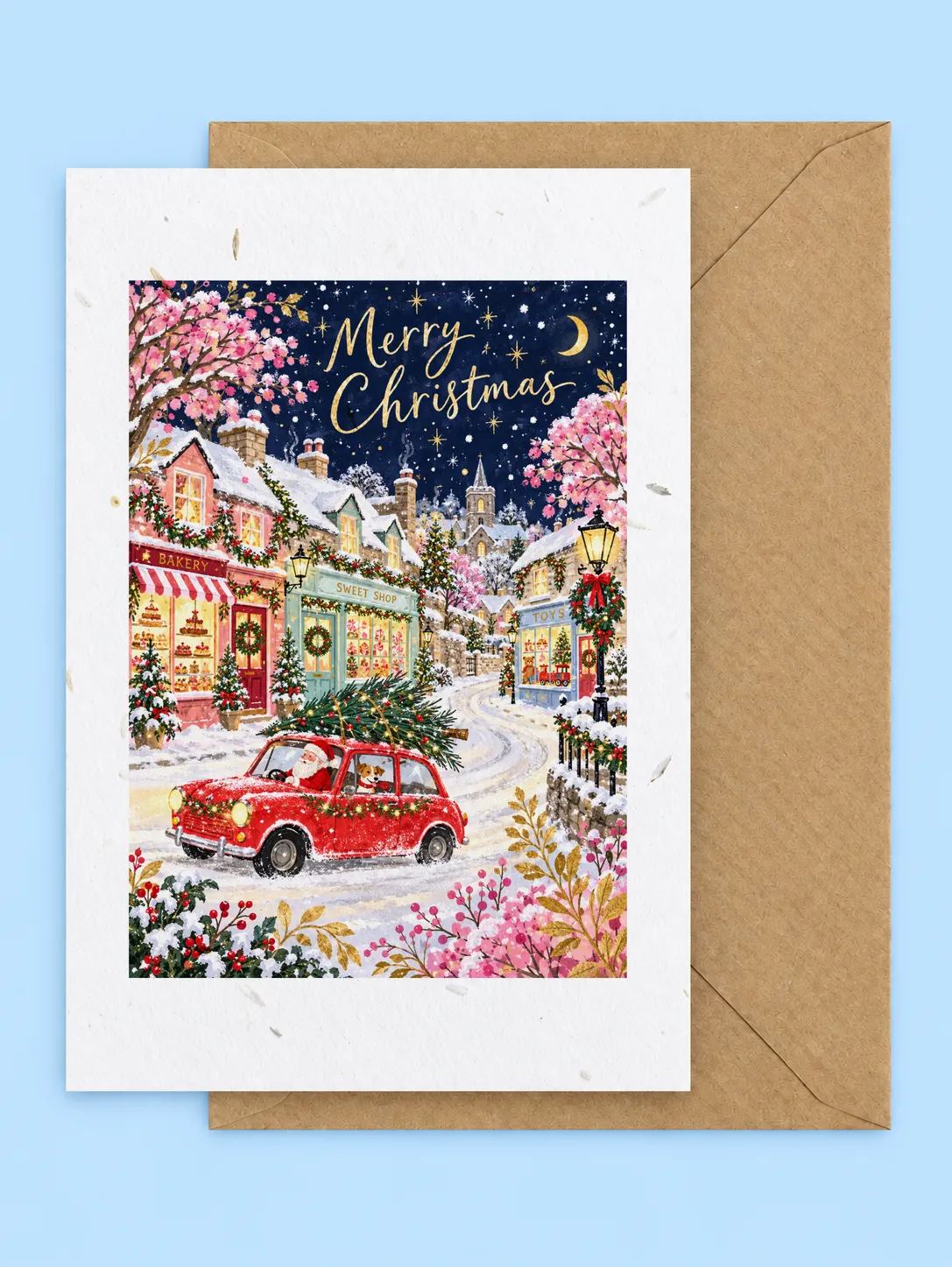 Snowy village street at night with a red car carrying a Christmas tree and glowing shop windows, seed paper Christmas card with a kraft envelope on a pale blue background