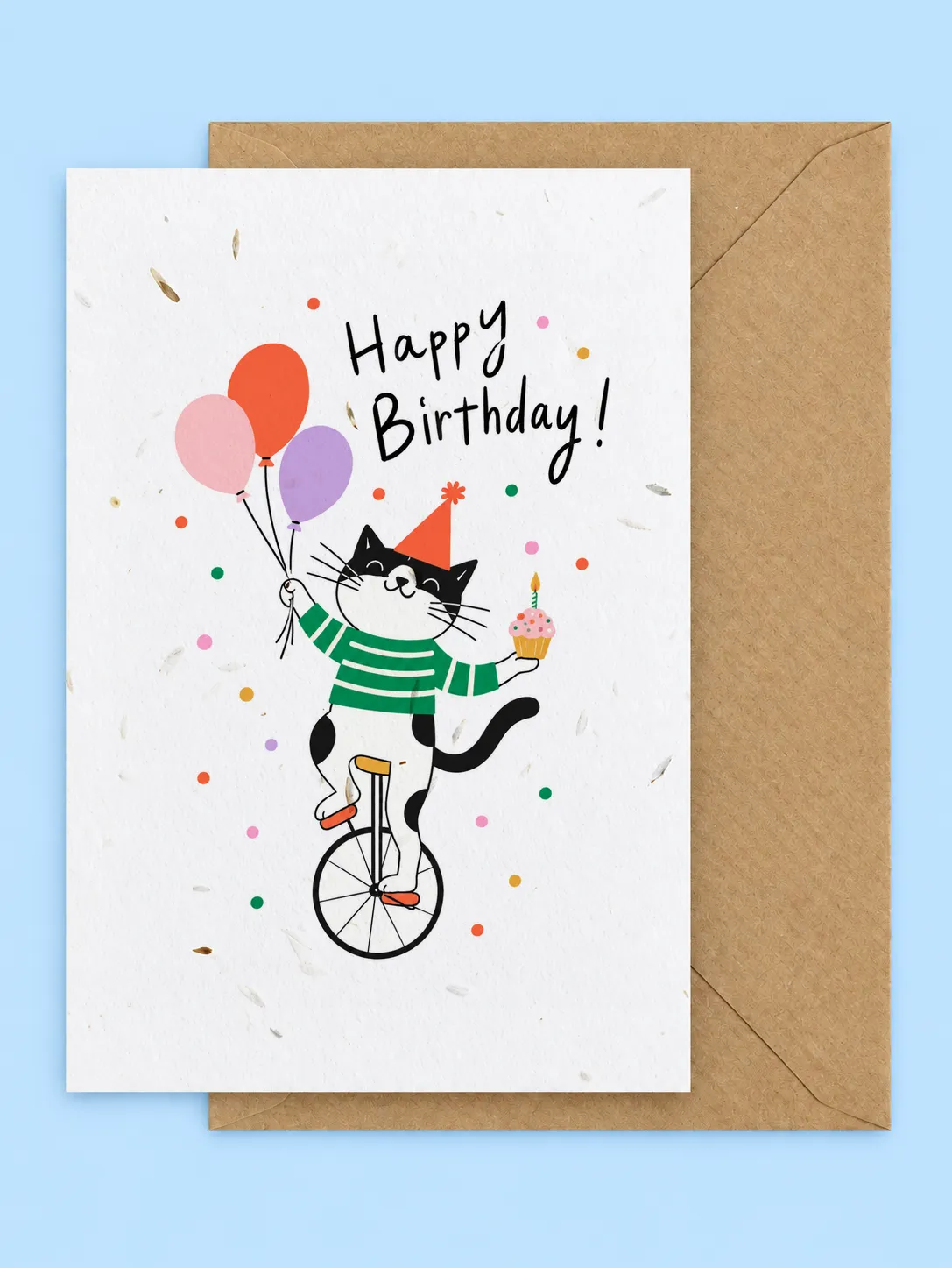 Black and white cat in a party hat riding a unicycle with balloons and a cupcake, seed paper birthday card with a kraft envelope on a pale blue background