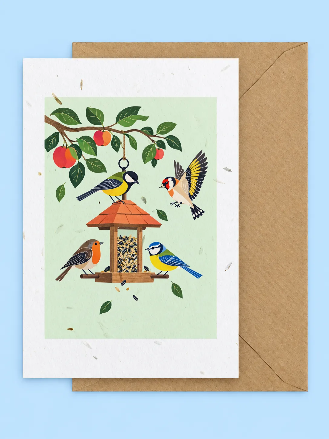 Blue tits, a great tit, a robin and a goldfinch around a bird feeder hanging from an apple branch, seed-filled greetings card with a kraft envelope on a pale blue background
