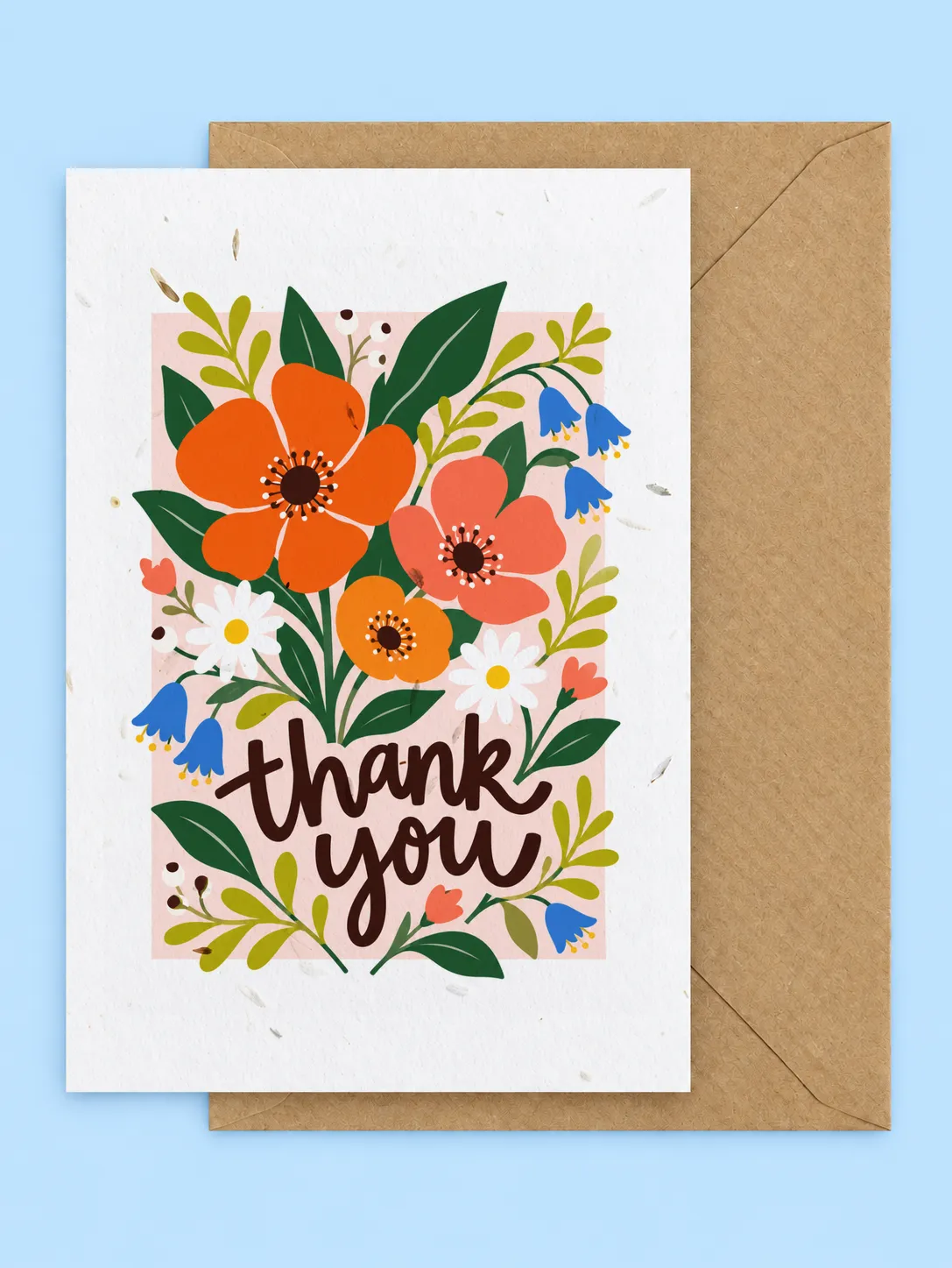 Bold orange flowers, bluebells and leaves around thank you lettering on a pink background, seed paper thank you card with a kraft envelope on a pale blue background