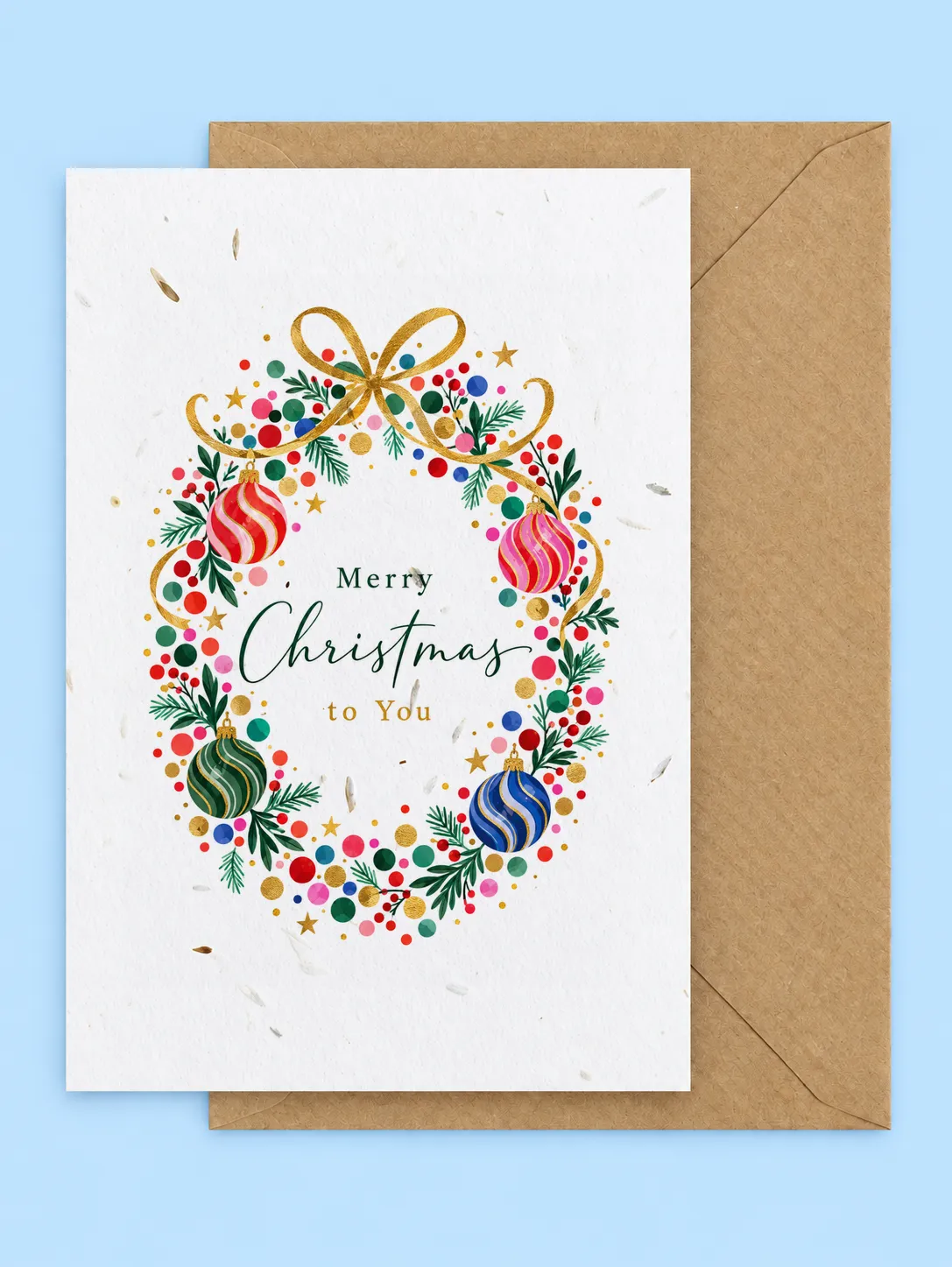 Christmas wreath of colourful baubles, berries and greenery with a gold bow, Merry Christmas to You plantable card with a kraft envelope on a pale blue background