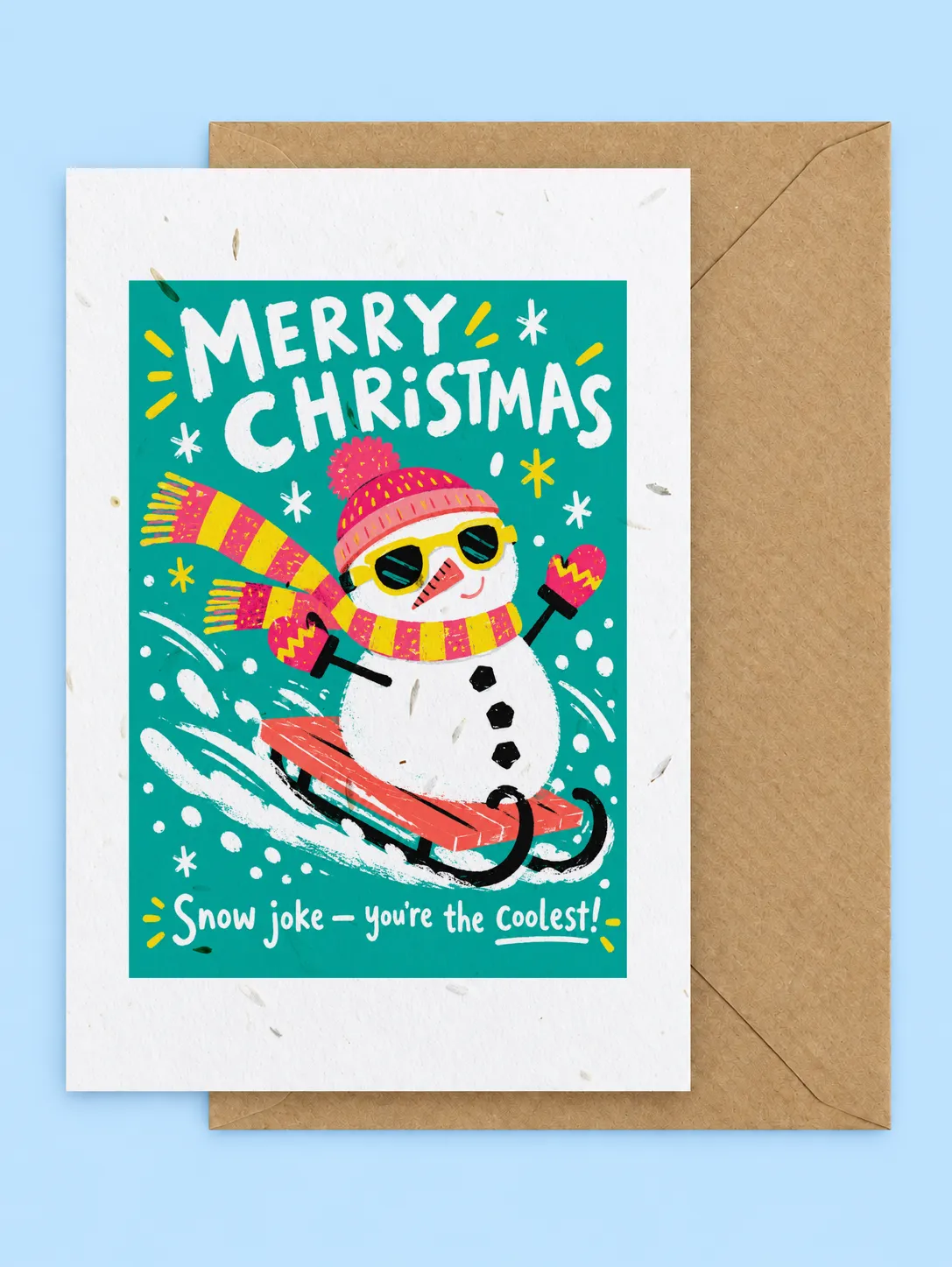 Snowman in sunglasses and a striped hat sledging on a teal background, Merry Christmas seed-filled card with a kraft envelope on a pale blue background